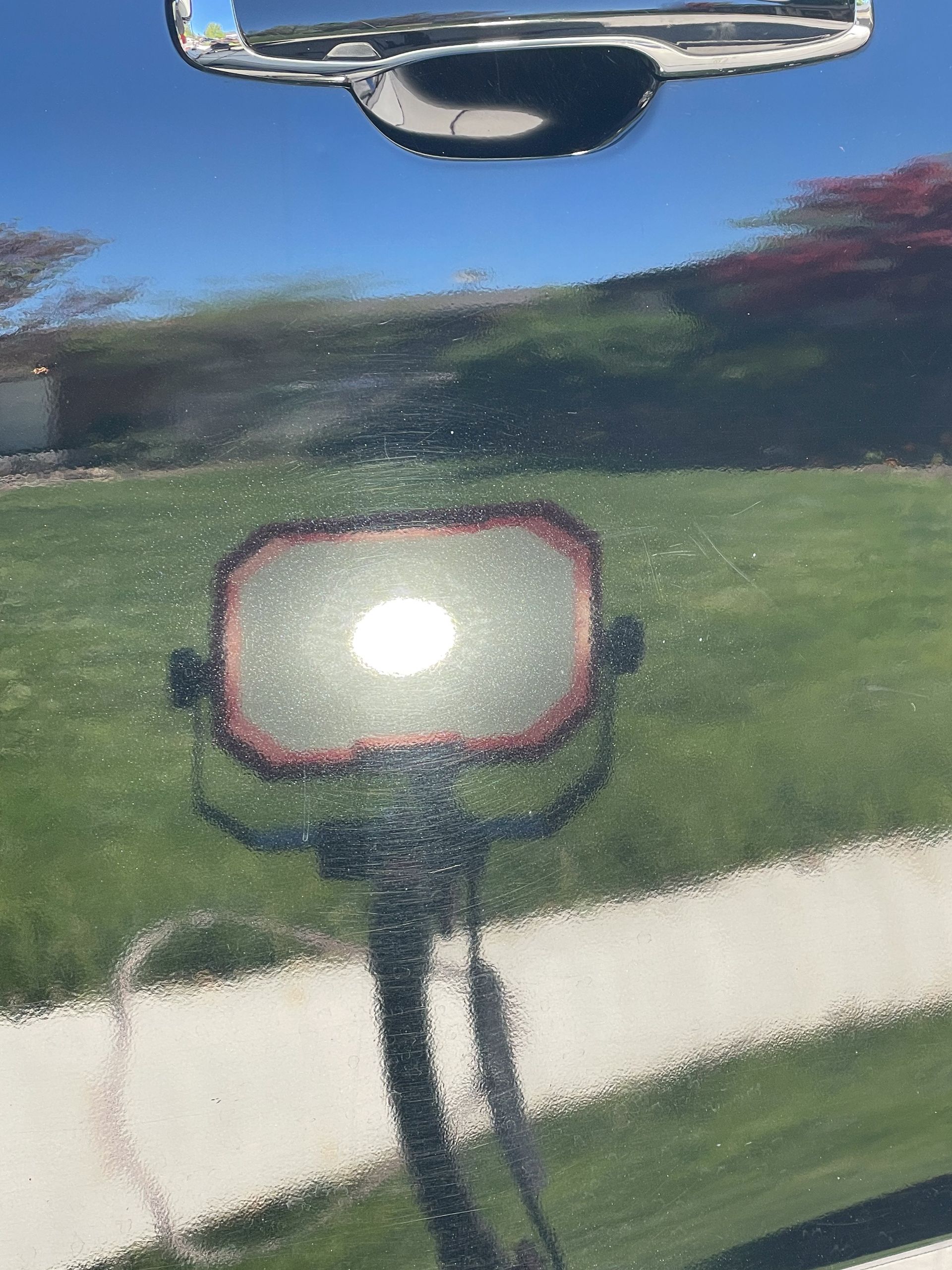 Reflection of a sunny day in polished black car hood; includes trees, sky, and spotlight.