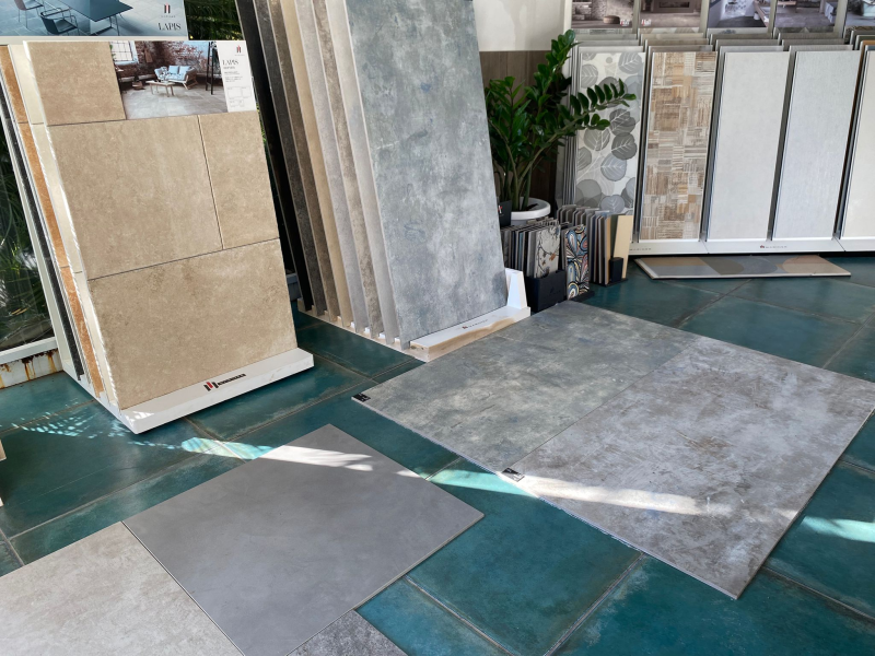Campioni di piastrelle esposti in uno showroom; vari colori e texture, dominano il grigio e il beige.