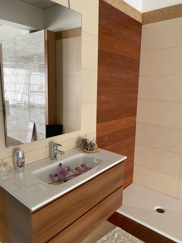 Bagno moderno con mobile lavabo in legno, specchio e doccia. La parete dietro la doccia è in legno, mentre le altre sono piastrellate beige.