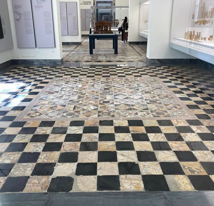 Pavimento a scacchiera in un museo, che conduce a un tavolo espositivo. Piastrelle quadrate chiare e scure.