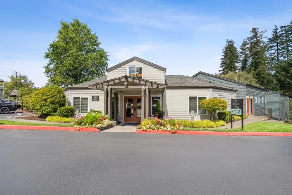 Gray building with a wooden trellis over the entrance and colorful flower beds.