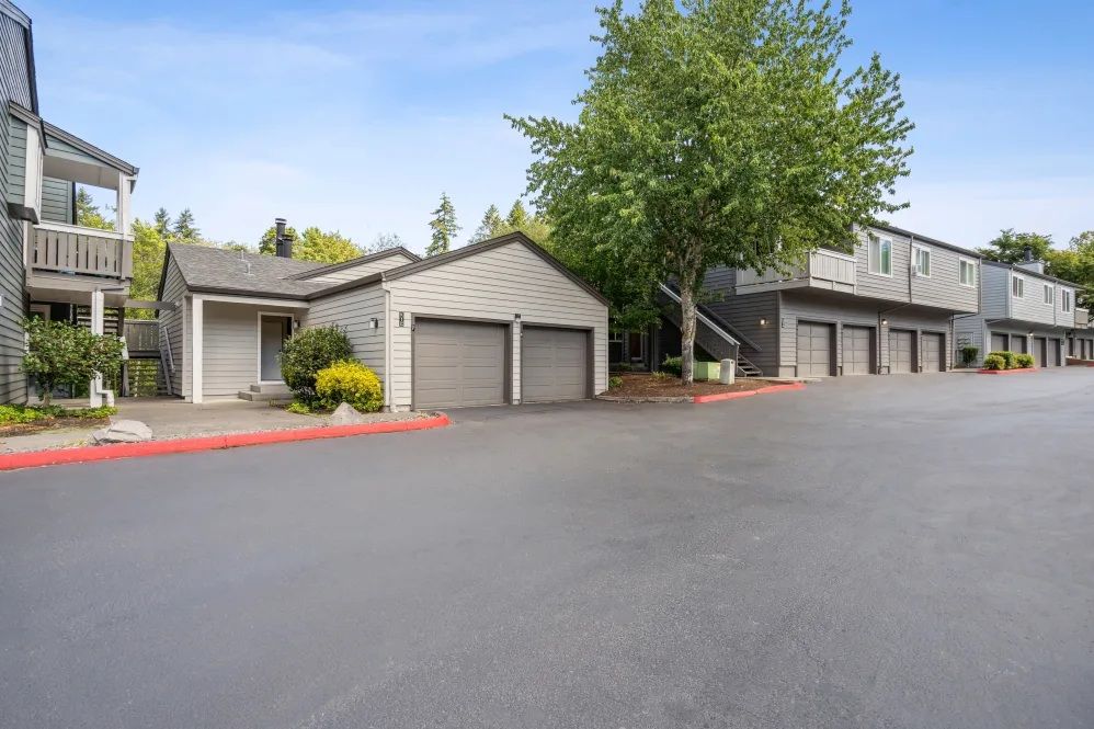 Apartment complex with garages and parking spaces; gray buildings, blue sky.