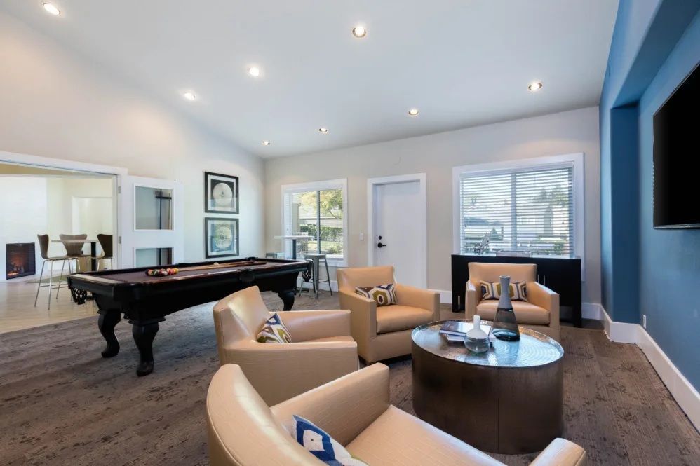 A game room with a pool table, armchairs, windows, and a large TV on a blue wall.