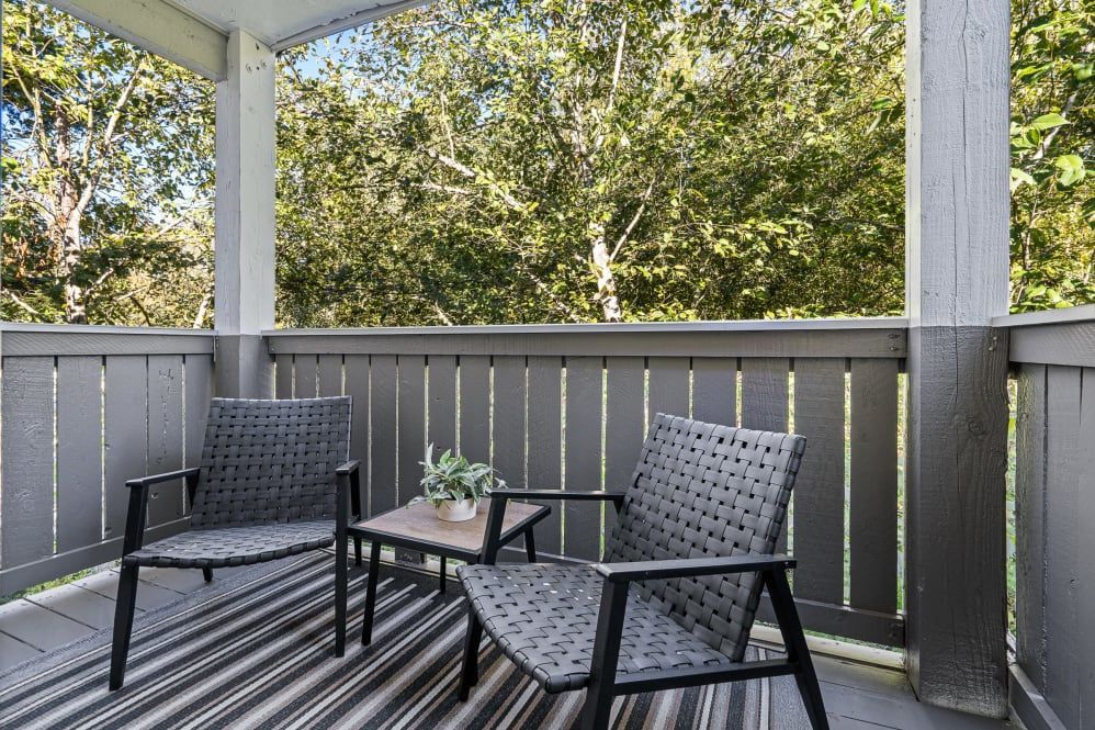 Two chairs and small table on a balcony overlooking trees. Gray and black furniture with a striped rug.