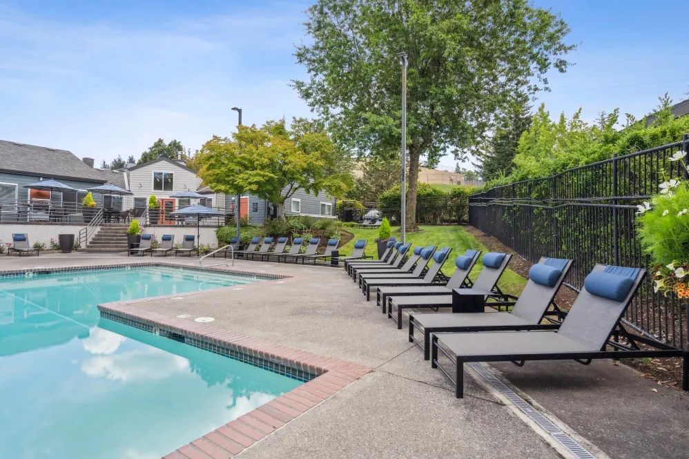 Swimming pool with lounge chairs on a sunny day. Building and trees in the background.