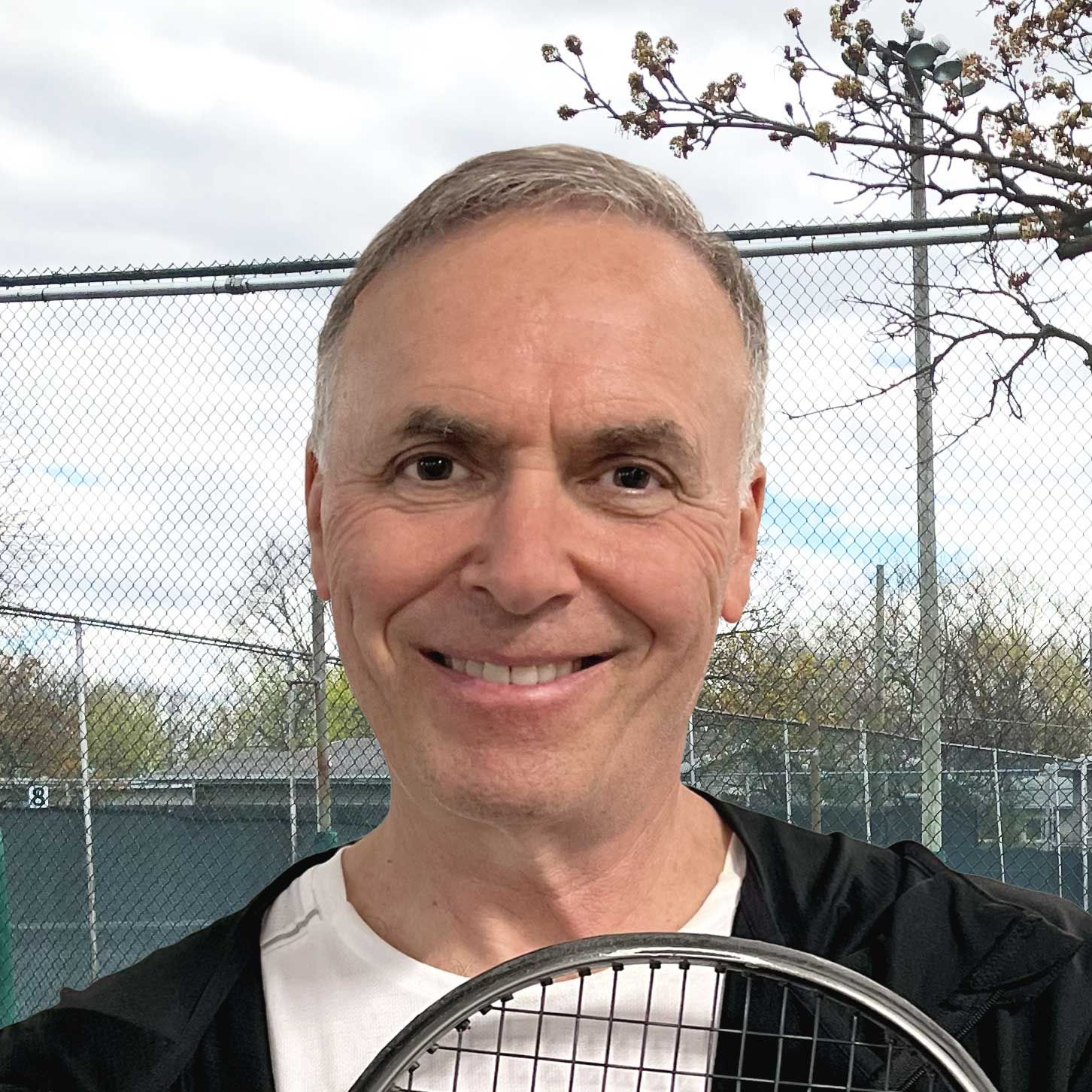 A man is smiling while holding a tennis racquet in front of a tennis court.