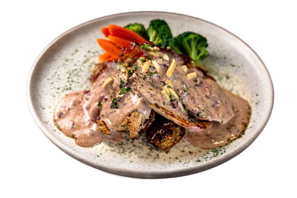 A plate of seared meat topped with creamy sauce, garnished with slivered almonds, sliced carrots, and steamed broccoli.