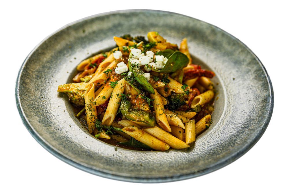 A plate of penne pasta tossed with vegetables and herbs, topped with a sprinkle of crumbled white cheese.