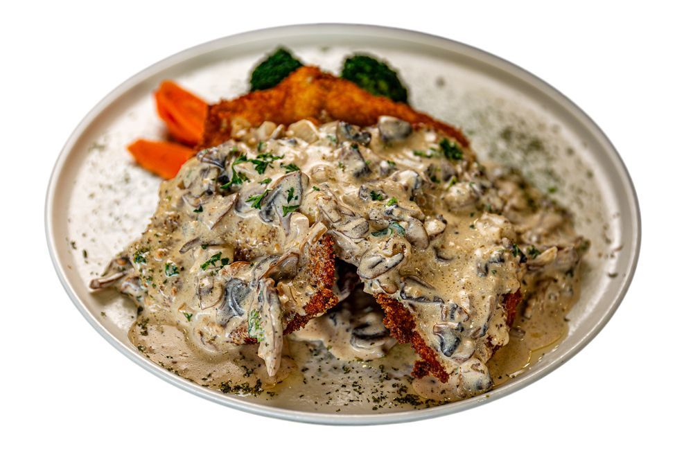 A breaded schnitzel topped with a creamy mushroom sauce, served on a white plate with small portions of cooked vegetables.