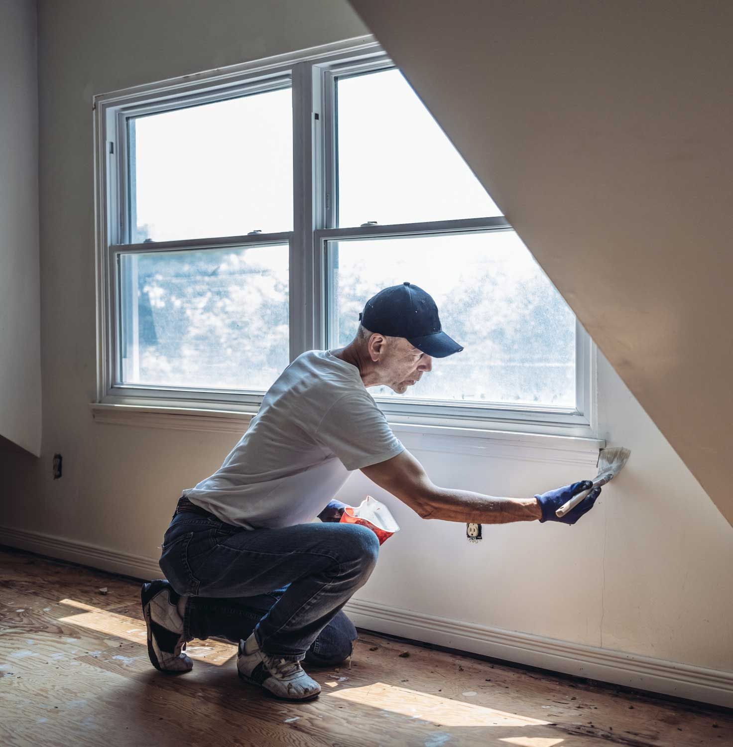 A man is kneeling down in front of a window while painting a wall.