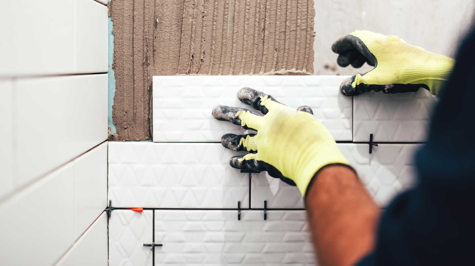 A man wearing yellow gloves is installing tiles on a wall.