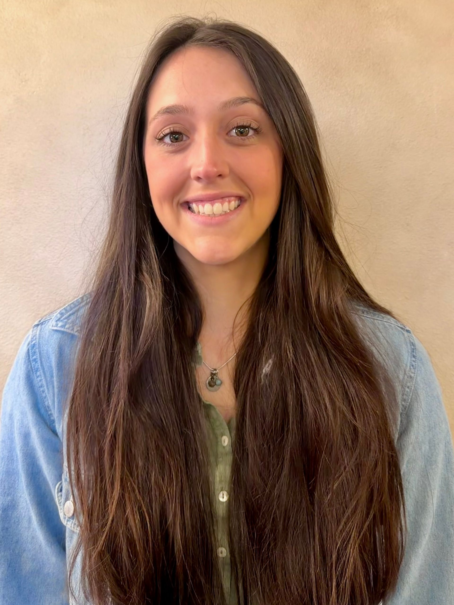 Smiling person with long dark hair wearing a denim shirt against a beige wall