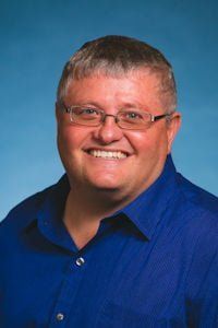 Smiling person in glasses wearing a blue shirt against a blue gradient background
