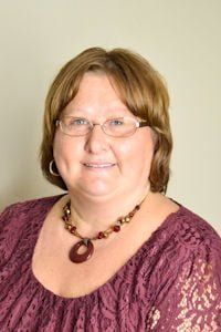 Smiling woman in glasses wearing a purple lace top and beaded necklace against a plain background