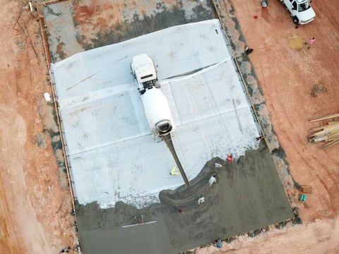 Concrete truck pouring cement into a construction site. Gray cement spreads on a rectangular surface surrounded by reddish soil.