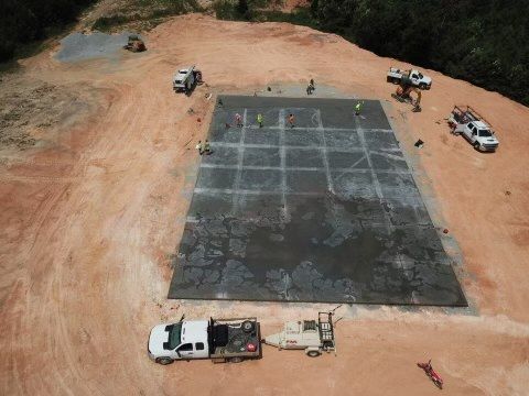 Construction site with workers on a large concrete pad, several trucks, and a small roller.