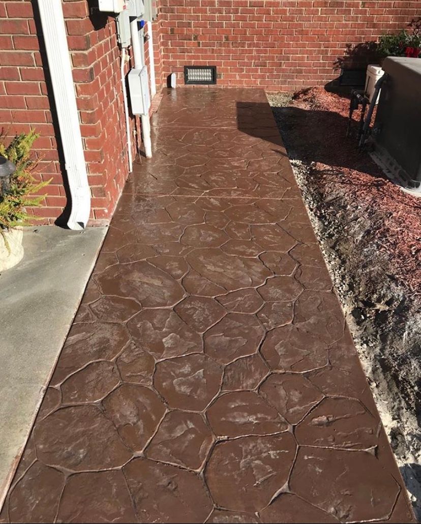 Brown, textured concrete walkway with a stone pattern next to a brick wall and a garden.