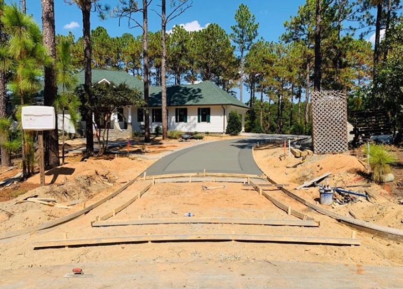 Driveway under construction with a newly paved section and wooden forms, leading to a white house with a green roof.