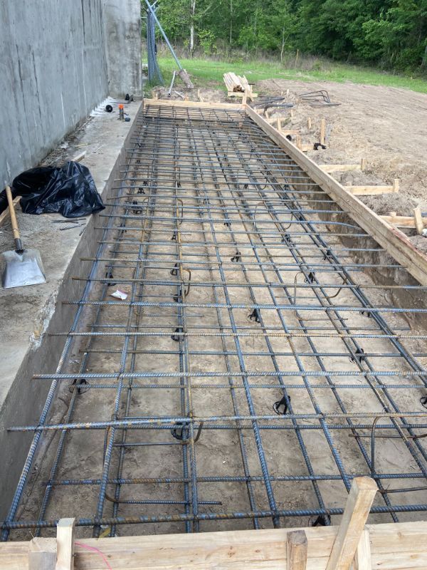 Rebar grid within wooden forms, prepared for concrete pouring outdoors near a concrete wall and wooded area.