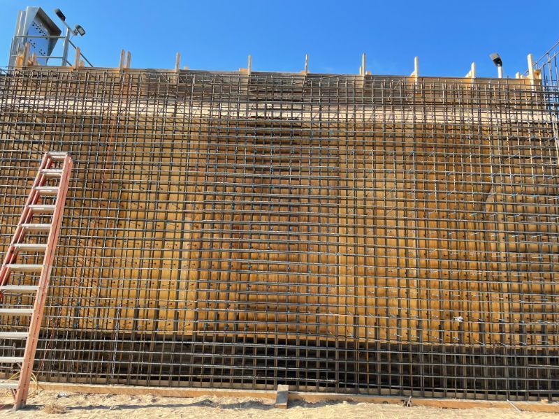 A large concrete wall under construction; rebar grid visible; red ladder leans against it. Clear blue sky.