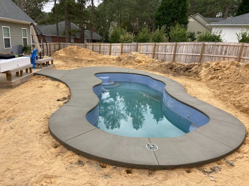 Concrete pool with water, surrounded by sand, wooden fence and house in the background.