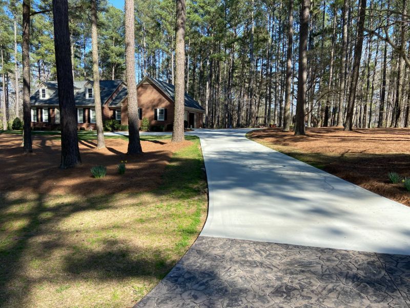 Concrete driveway curves toward a brick house in a wooded area. Sunny day.