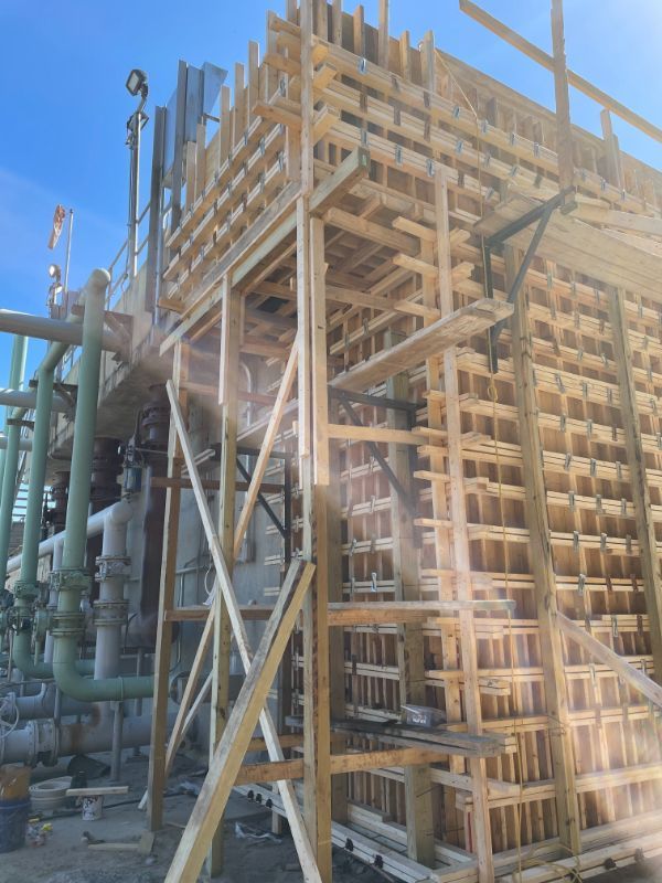 Wooden scaffolding and concrete formwork in a construction setting, next to industrial pipes and equipment.