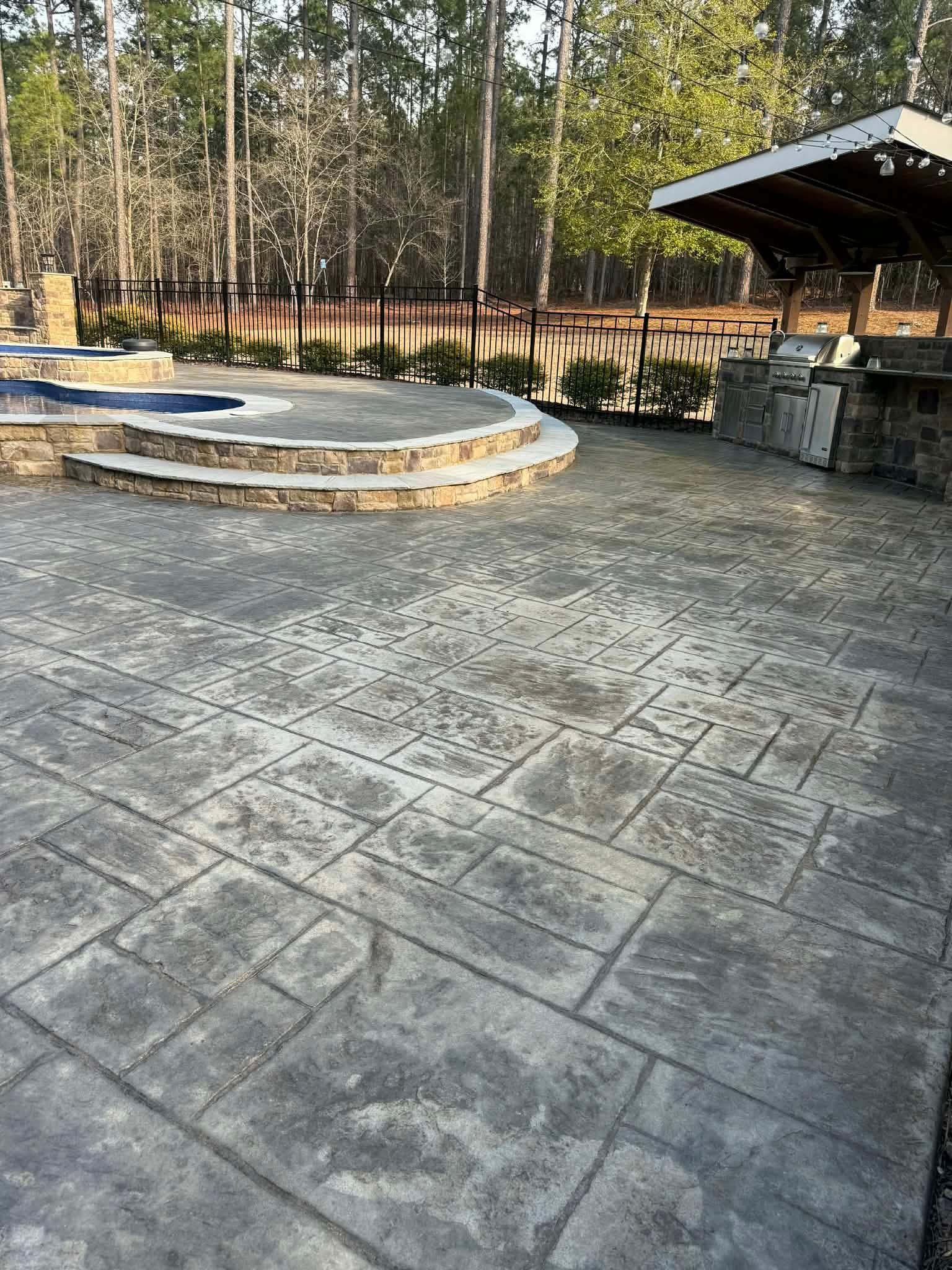 Black stamped concrete driveway leads to a white house with a two-car garage. Brown grass and trees surround.