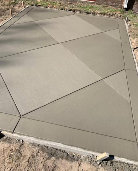 Concrete patio with a blue rectangular pool surrounded by trees. White chairs stacked on the right. Concrete patio surrounds a blue rectangular pool; trees and a building are in the background.
