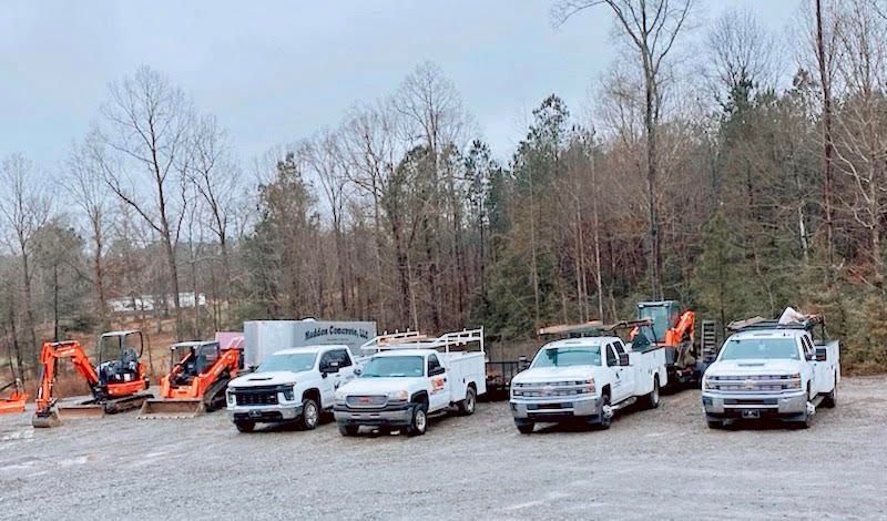 Construction vehicles parked on gravel, with trees in the background under an overcast sky.