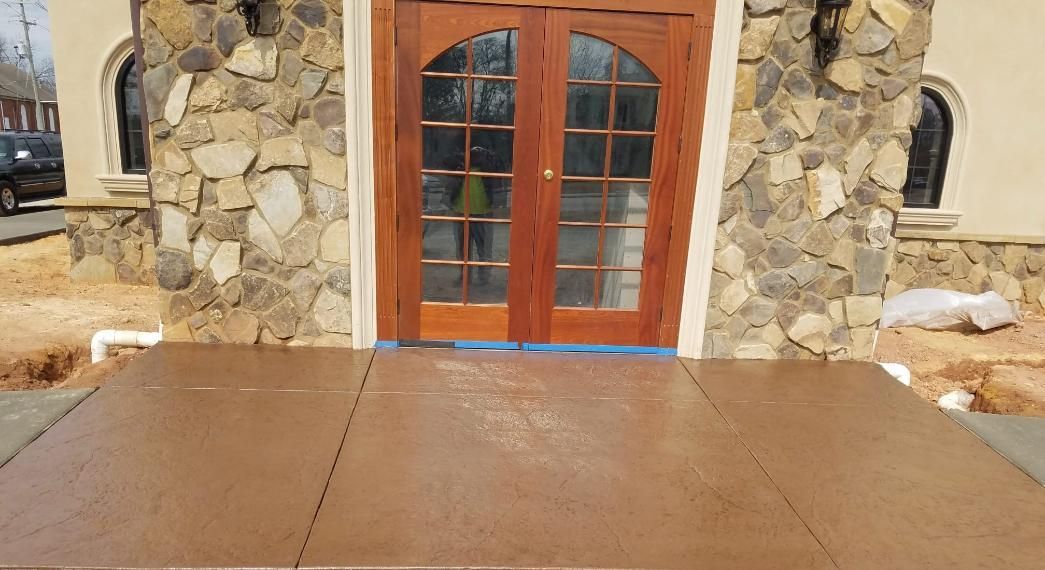 Brown, textured concrete patio in front of a building with stone columns and a treeline in the background. Stamped concrete patio in front of a building with stone columns. Brown and tan colors.