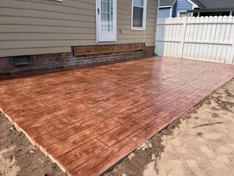 Stamped concrete walkway in brown and gray tones, curving in yard next to dirt. Stamped concrete walkway, brown and gray stone pattern, curving through a dirt area.