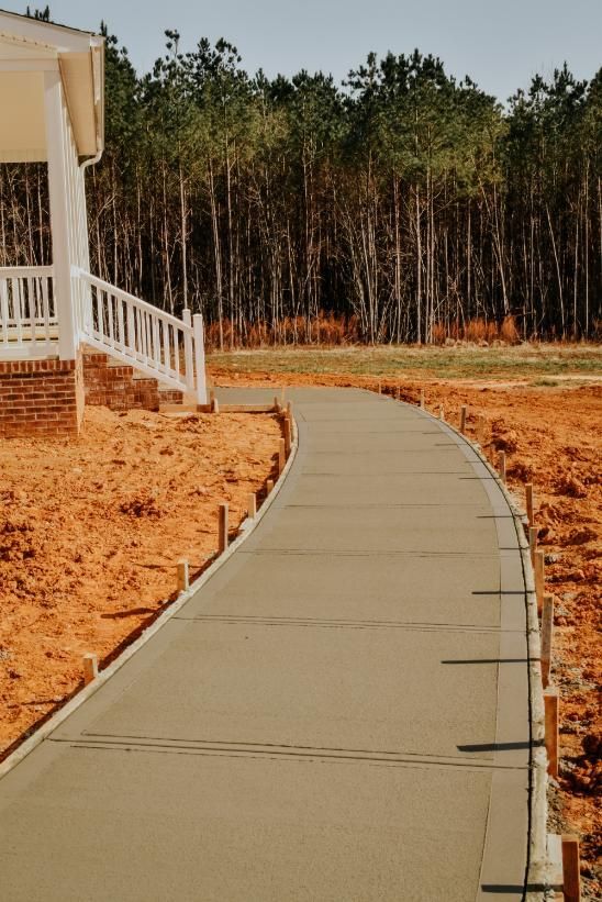 Newly poured concrete walkway leading to a house, flanked by wooden forms on a construction site. Newly poured concrete walkway curves towards a forest, next to a house with a porch.