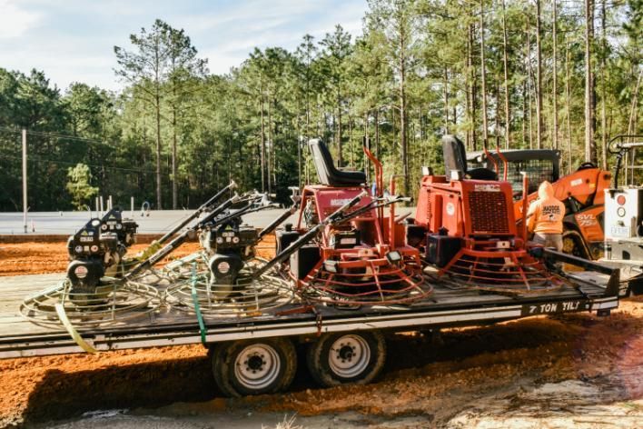 Red concrete finishing machine on a trailer at a construction site. Man in orange vest. Forest in the background.