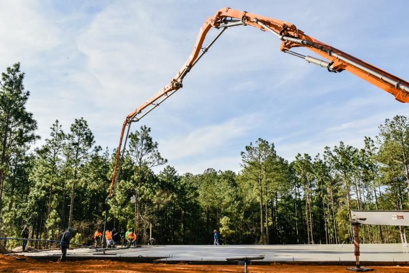 Concrete pump truck pouring concrete slab on construction site surrounded by trees.