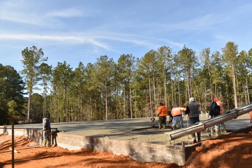 Construction workers pouring concrete foundation; outdoor setting with trees and blue sky.