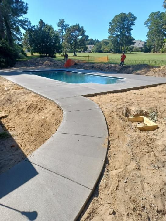 Finished wood-look stamped concrete patio, red-brown, next to a house with beige siding and a white fence. Stamped concrete patio with wood-grain pattern and reddish-brown stain next to a house with a white fence.
