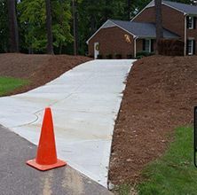 Orange traffic cone on a concrete driveway leading to a brick house. Mulch borders the driveway.