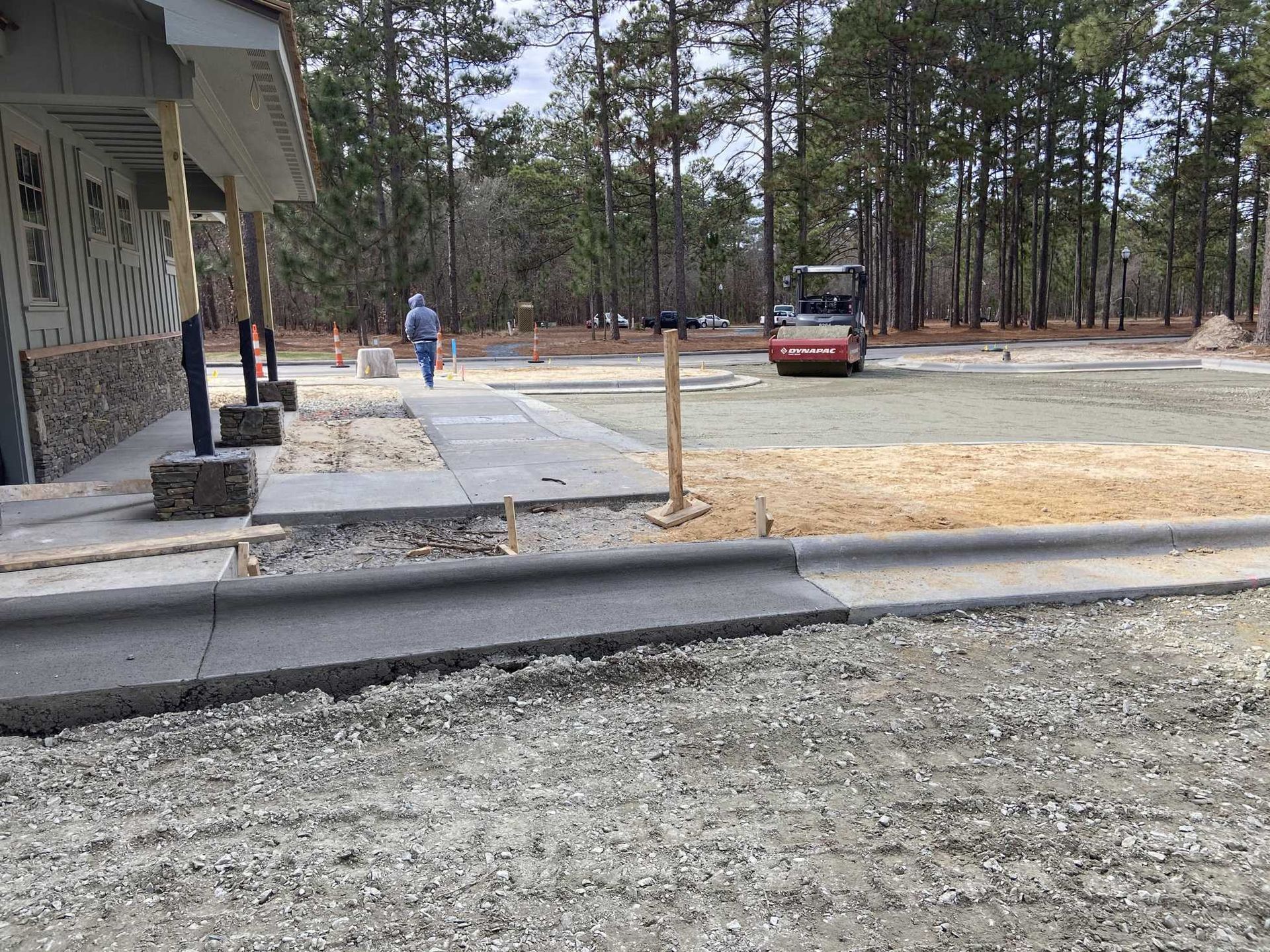 Construction site: Paved walkway, curb, gravel, small roller, person walking towards a restroom in a forest.