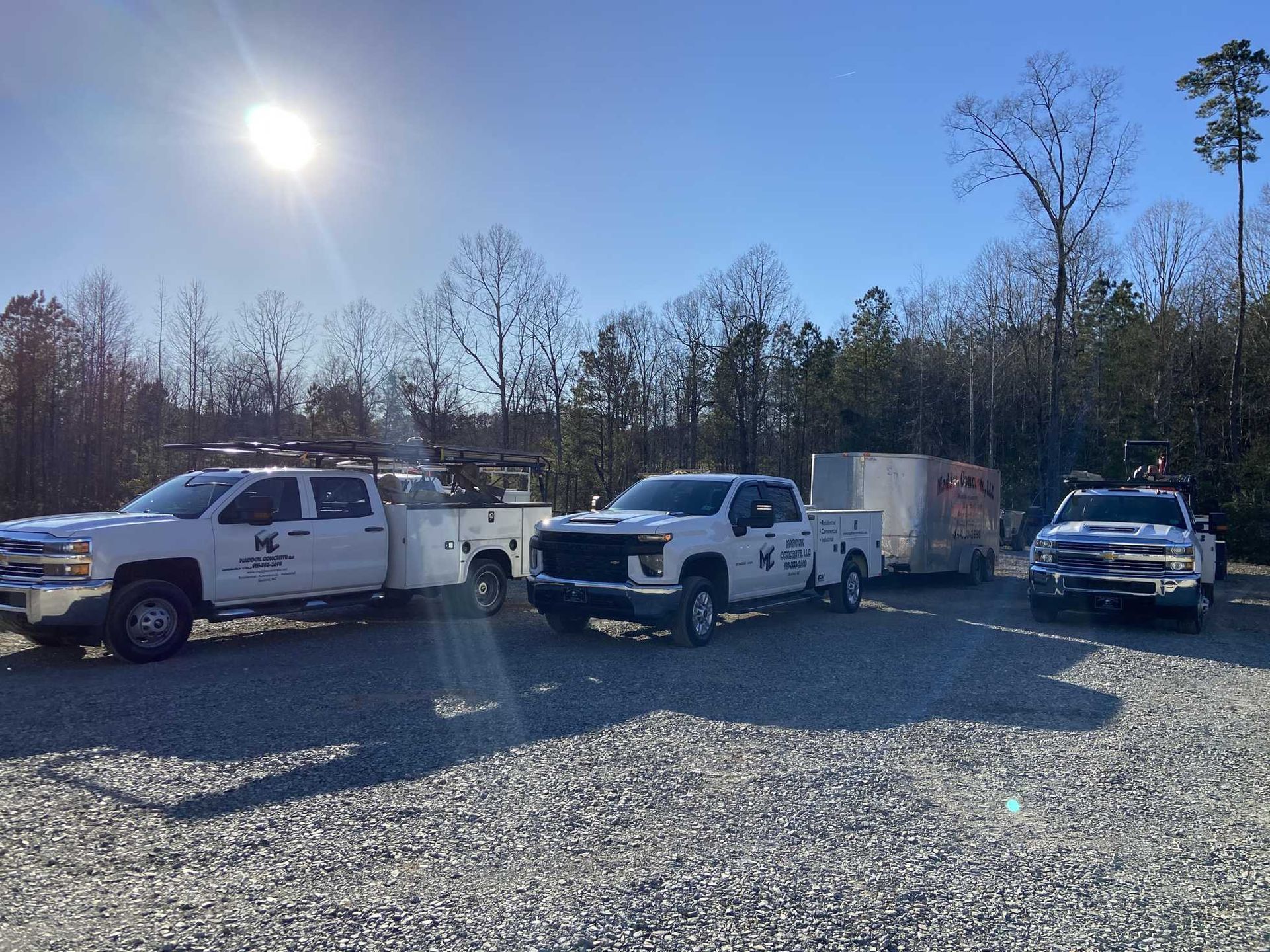 Three white work trucks parked on gravel, sunny day. One towing a trailer.