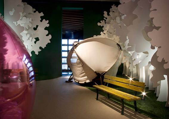 stanza con una panchina, prato finto con alberi di cartone di color bianco, un amaca moderna da giardino e sulla sinistra si intravede una palla rosa