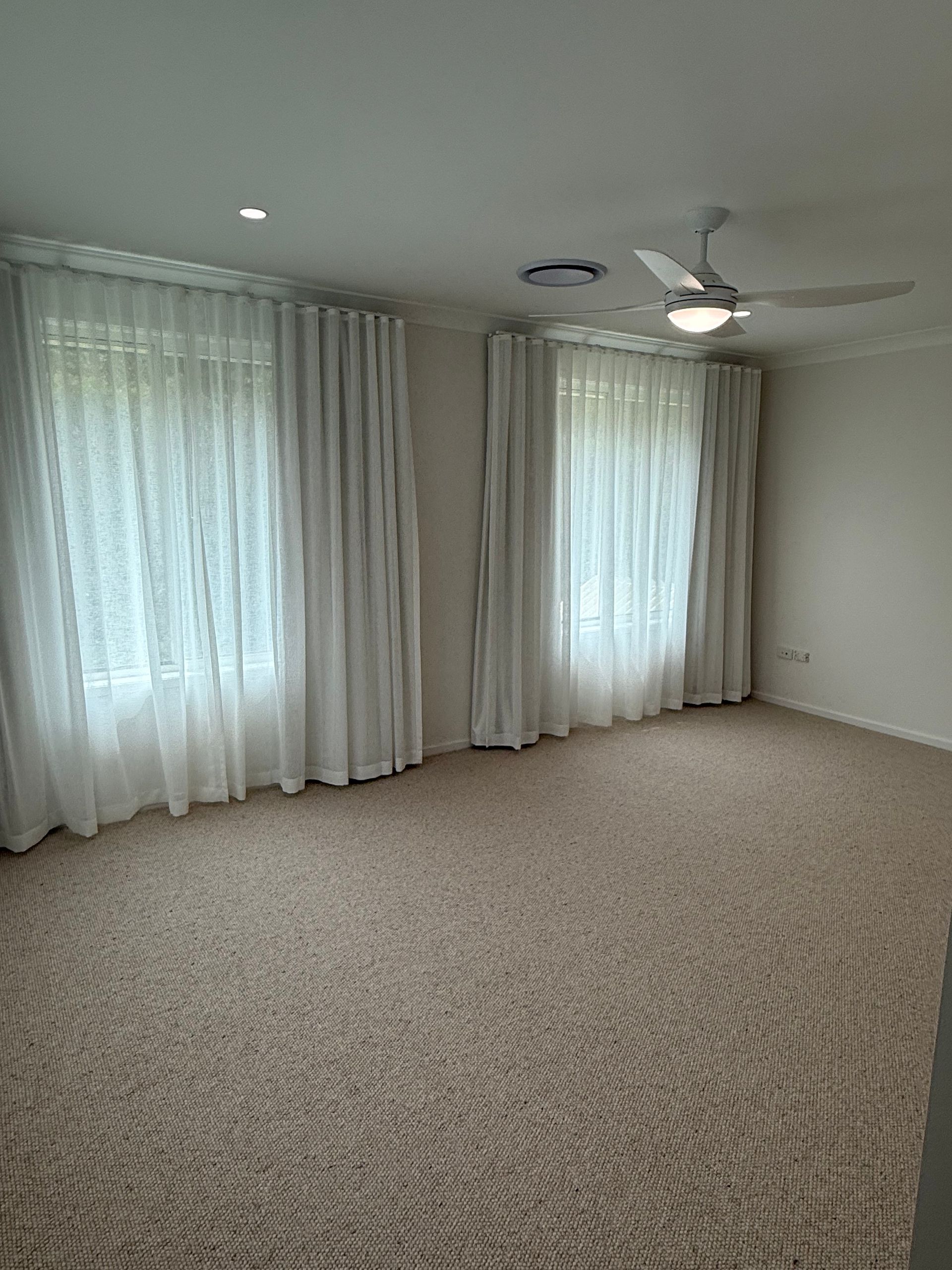 Empty room with two windows, sheer white curtains, beige carpet, and a ceiling fan.
