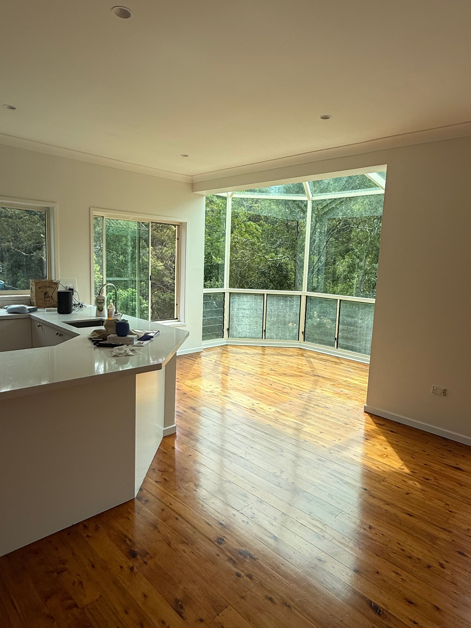 Bright kitchen with wooden floor, white countertop, and large windows looking out to trees.