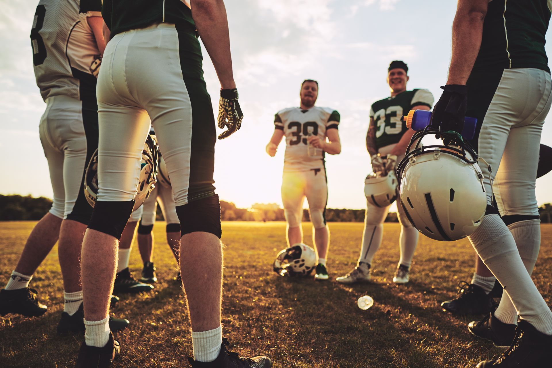 A group of American football players in uniforms standing in a huddle on a field during a golden sunset.