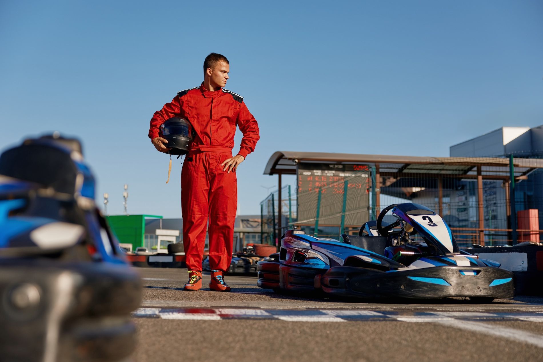 A racer in a red suit stands holding a helmet next to a go-kart on a track under a clear blue sky.