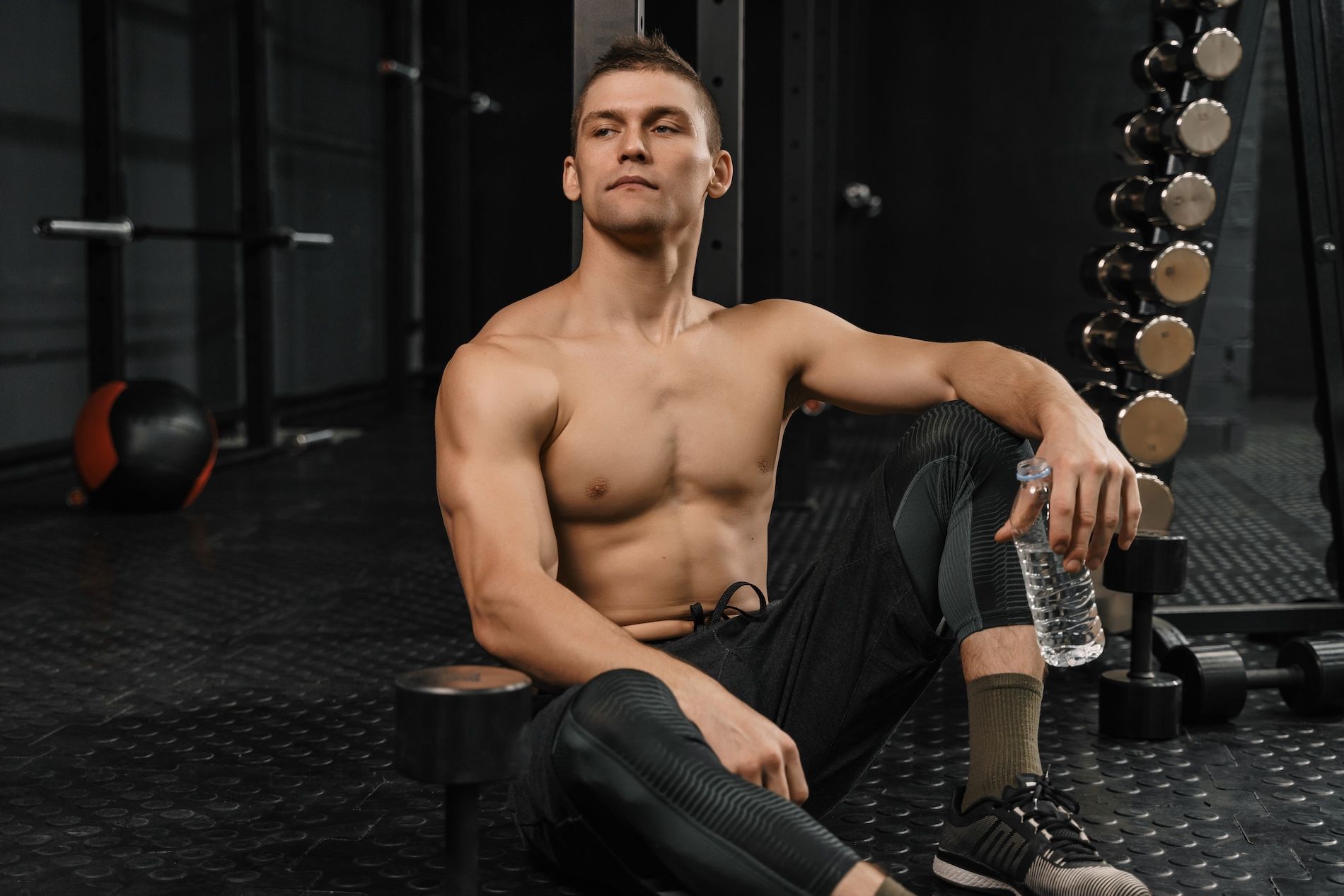 A shirtless person resting in a gym, holding a water bottle while sitting on the floor near a dumbbell rack.