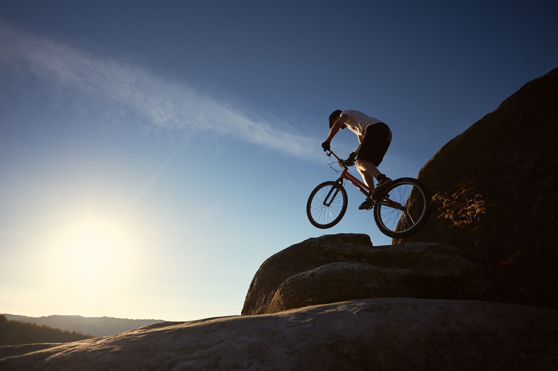 A cyclist performs a stunt on a mountain bike atop a rocky peak silhouetted against a bright, clear sky.
