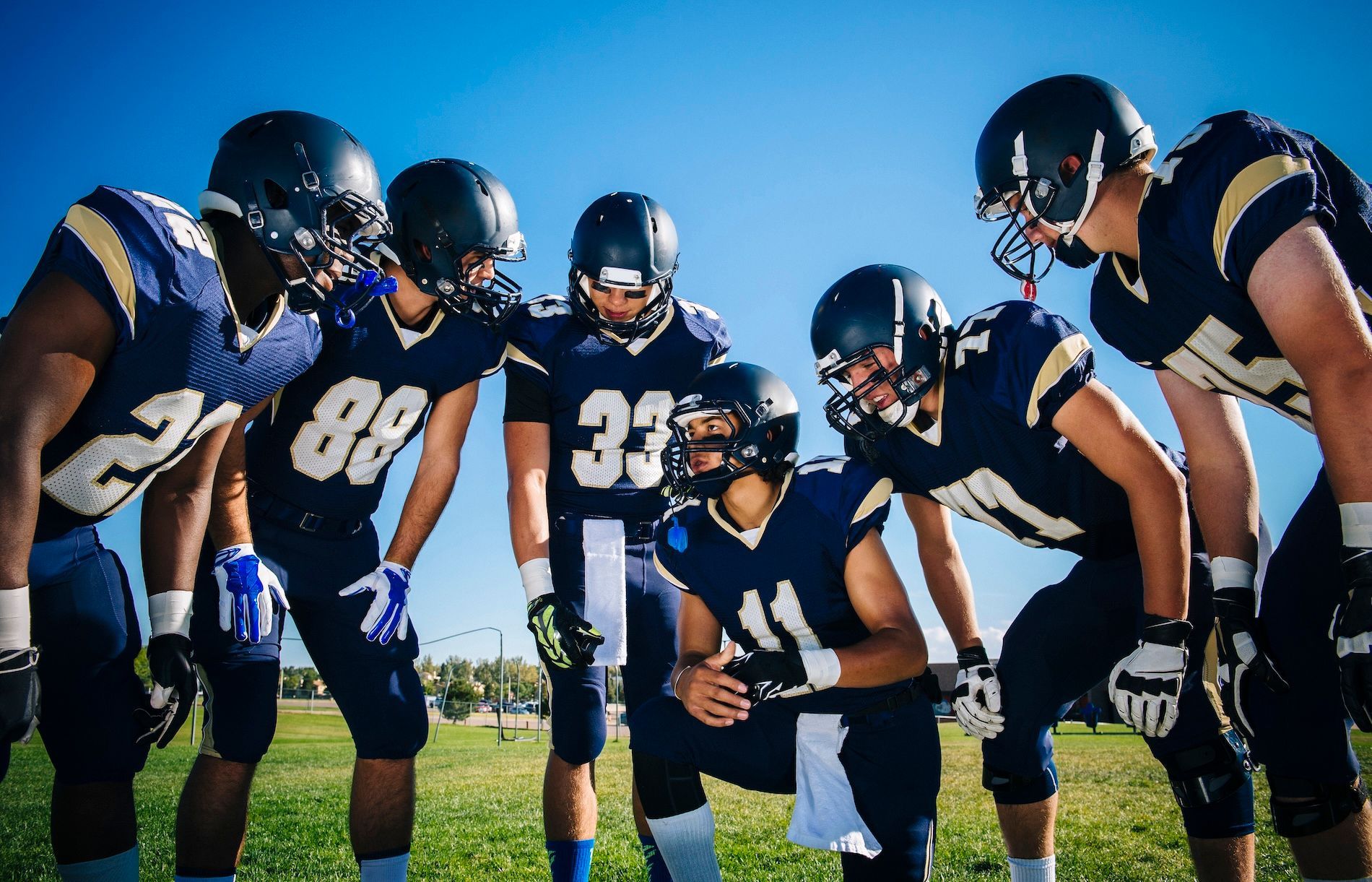 A group of football players in navy uniforms huddled together on a green field under a clear blue sky.
