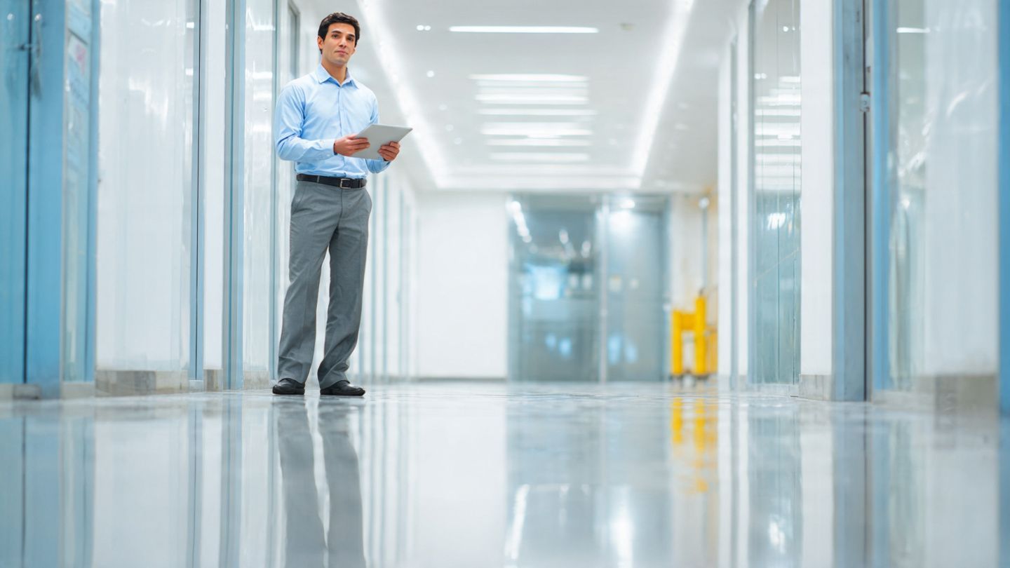 A professional standing in a bright, modern office corridor, holding a tablet while looking toward the camera.
