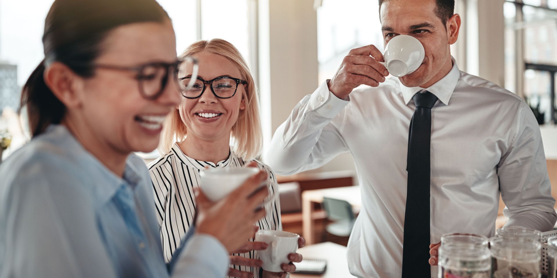 Three colleagues laughing and drinking coffee in an office, one wearing a tie.